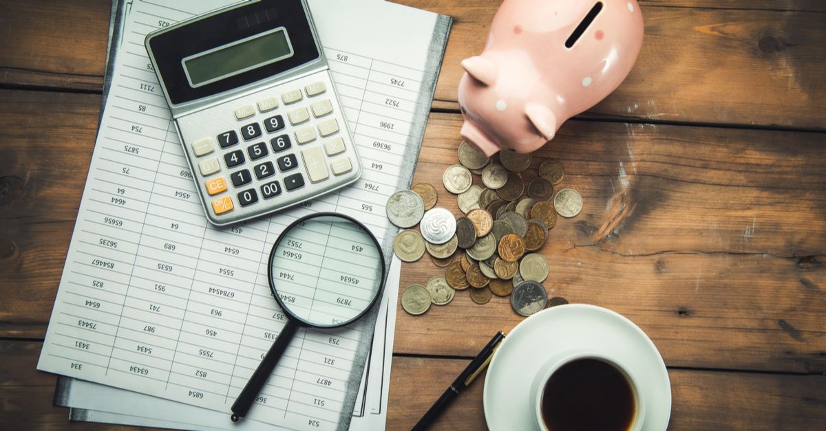 A desk with paperwork, a calculator, a magnifying glass, a bunch of coins, a cup of coffee, and a piggy bank on it