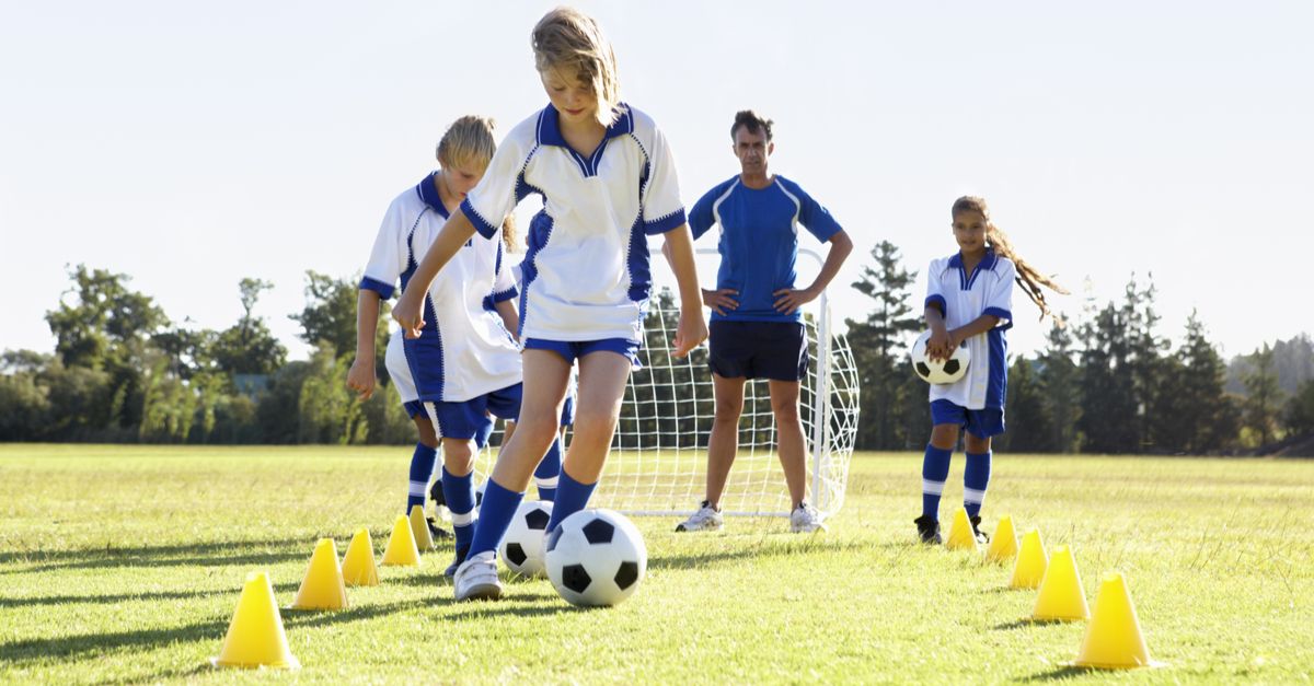 A photo of four children at football trials, dribbling the ball through small yellow cones while the coach watches